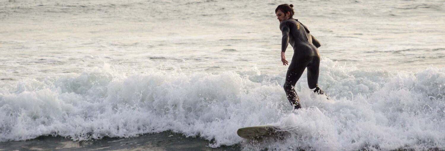 Uma pessoa em uma roupa de mergulho surfando em pequenas ondas perto da costa, com o oceano ao fundo no Curso de surfe na Praia Makaha