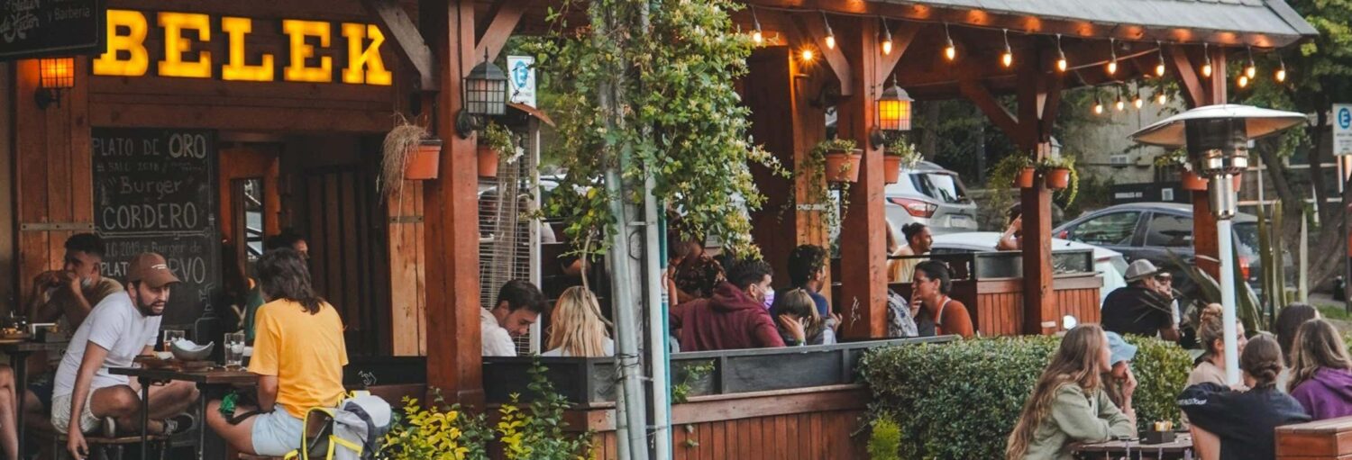As pessoas se sentam ao ar livre em mesas de madeira sob luzes de corda em um restaurante com uma grande placa "BELEK" na parede na Degustação de cerveja artesanal no Belek