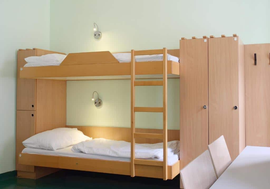 Um quarto no Jugendherberge Wien - Myrthengasse com beliches de madeira, roupa de cama branca, dois guarda-roupas e duas cadeiras contra uma parede branca, onde fica em Viena.