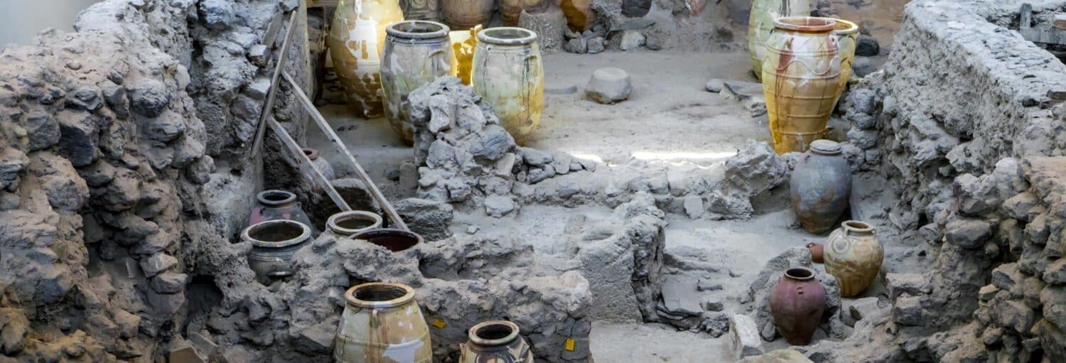 Grandes jarros de cerâmica antigos e ruínas de pedra são mostrados em um local de escavação arqueológica com solo empoeirado e paredes rochosas. A imagem ilustra o post sobre Ingresso do sítio arqueológico de Akrotiri.