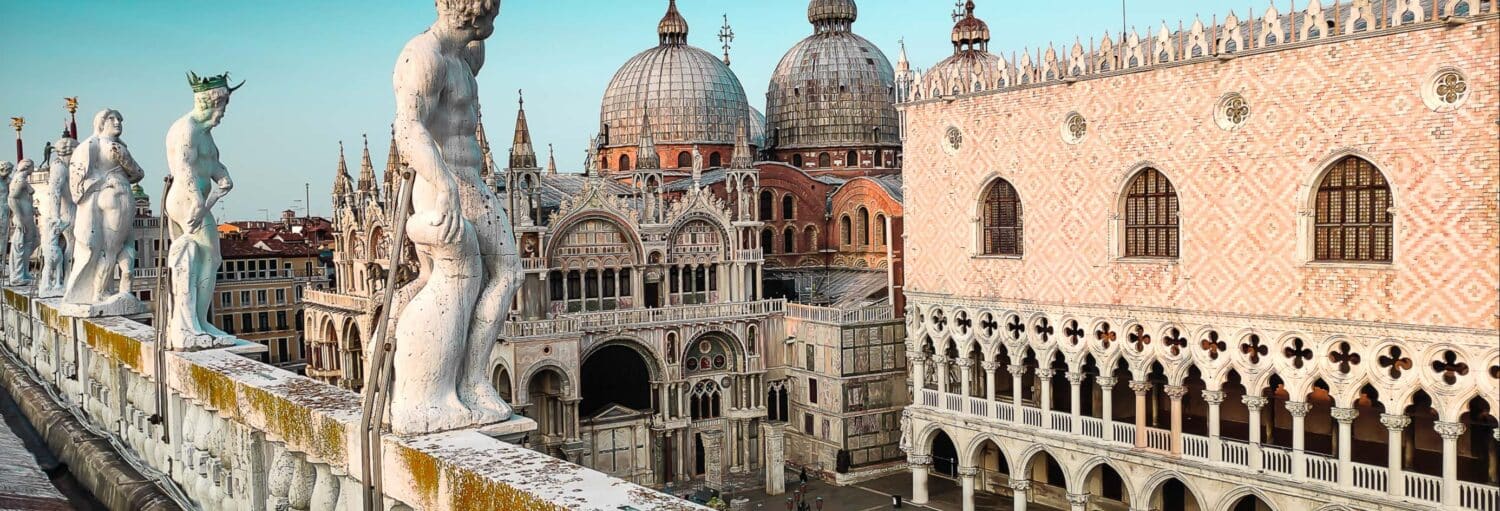 Estátuas alinham-se em um telhado com edifícios abobadados e ornamentados, incluindo a Basílica de São Marcos, em Veneza, Itália. A imagem ilustra o post sobre Ingresso da Basílica de São Marcos.