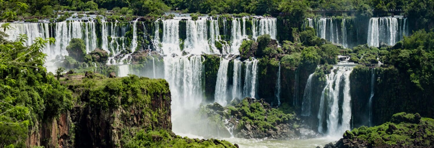 Ampla vista das Cataratas do Iguaçu com várias cachoeiras caindo em cascata sobre penhascos rochosos cercados por uma vegetação verde exuberante. - Ingresso do lado brasileiro das Cataratas do Iguaçu