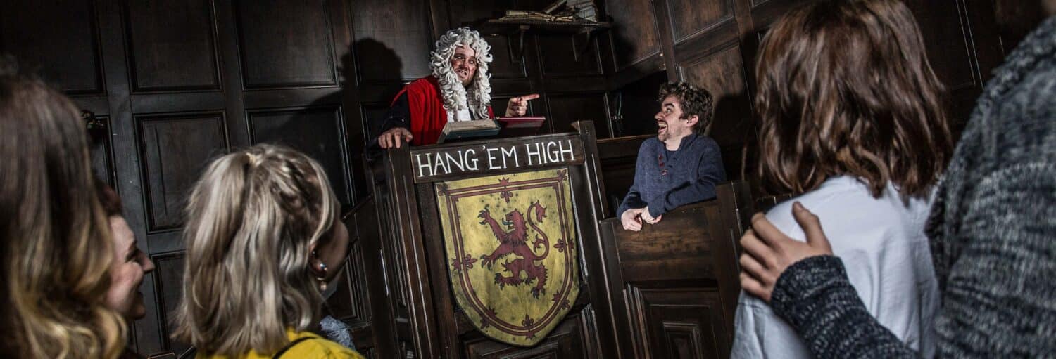 Um juiz com peruca branca e túnica vermelha aponta para um homem sorridente em uma sala de audiências com uma placa que diz "Hang 'Em High". A foto é capa do post do Ingresso do The Edinburgh Dungeon.