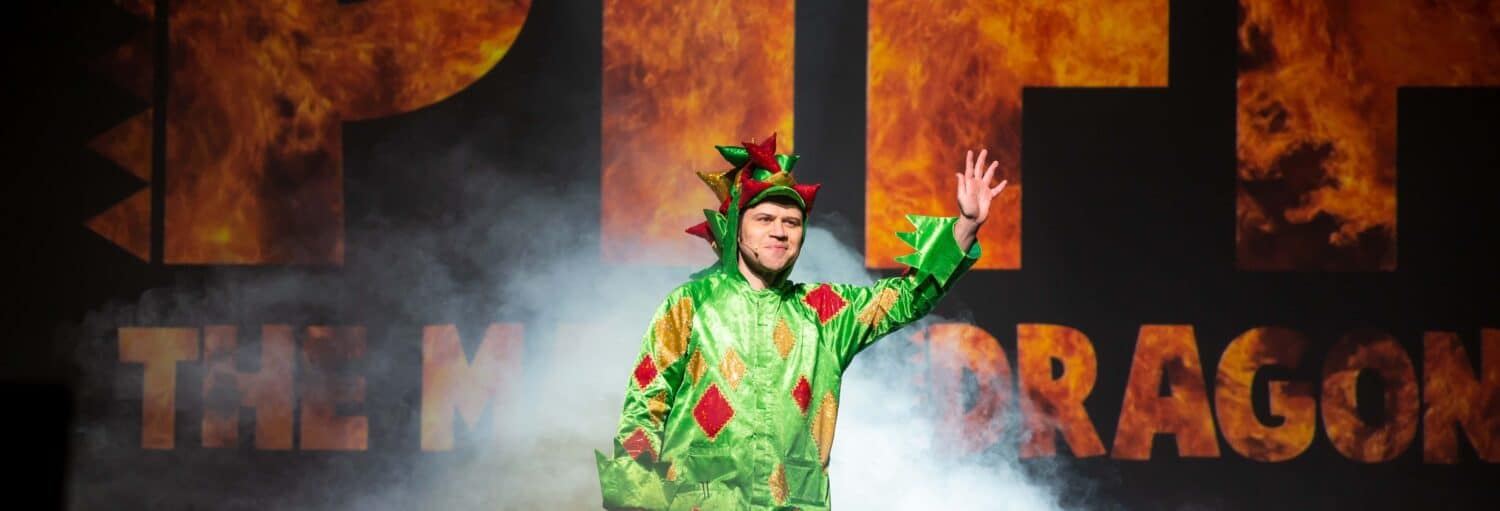 Uma pessoa com uma fantasia de dragão verde está no palco, acenando, com efeitos de fumaça e texto de fundo com tema de fogo atrás dela. Imagem para ilustrar post sobre ingresso do espetáculo Piff the Magic Dragon.