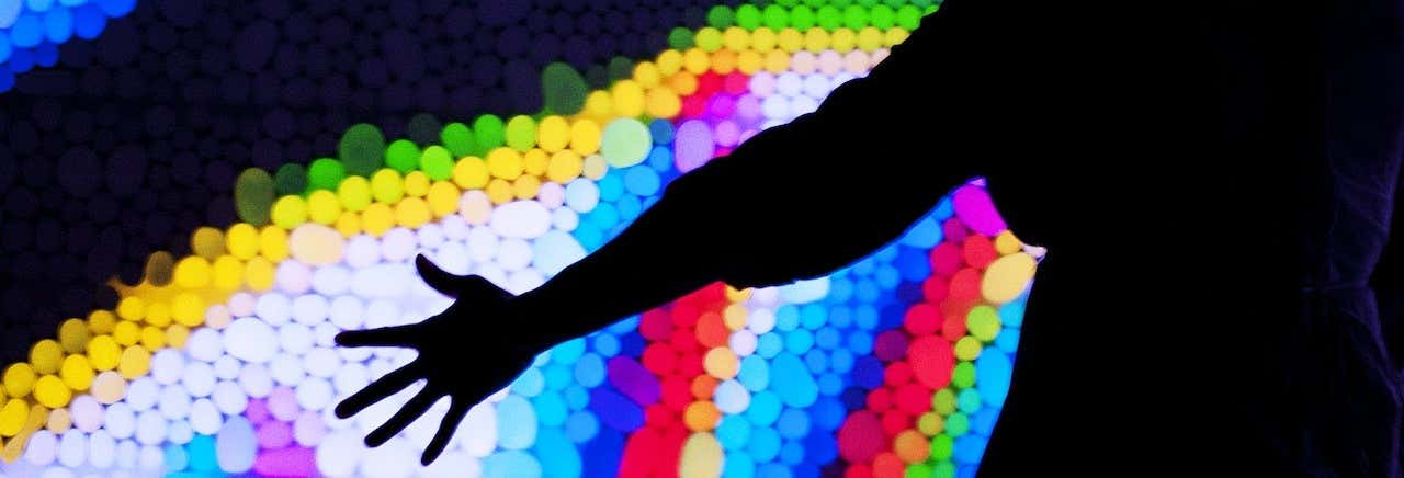 Silhueta de uma pessoa, com seu braço em foco. Sua mão passa por uma parede cheia de bolas coloridas. Ingresso do Exploratorium