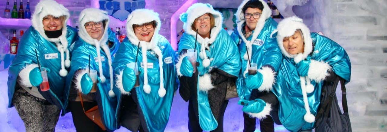 Seis pessoas com capas azuis com capuz estão juntas em um bar de gelo, segurando bebidas e sorrindo para a câmera. Imagem para ilustrar post sobre o ingresso do Ice Bar.