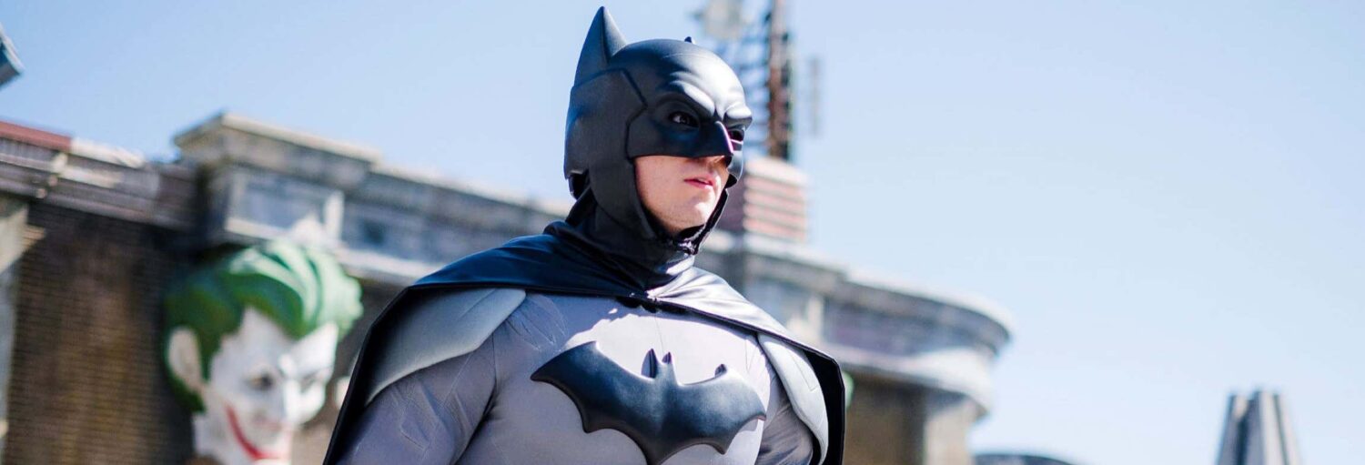 Uma pessoa em uma fantasia de Batman está do lado de fora, com prédios e o rosto do Coringa visíveis ao fundo. A imagem ilustra o post sobre Ingresso do Parque Warner.