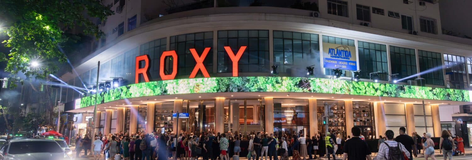 Uma multidão está do lado de fora do cinema Roxy à noite, com grandes letras vermelhas e luzes brilhantes iluminando a entrada. A imagem ilustra o post sobre Ingresso do Roxy Dinner Show com jantar.