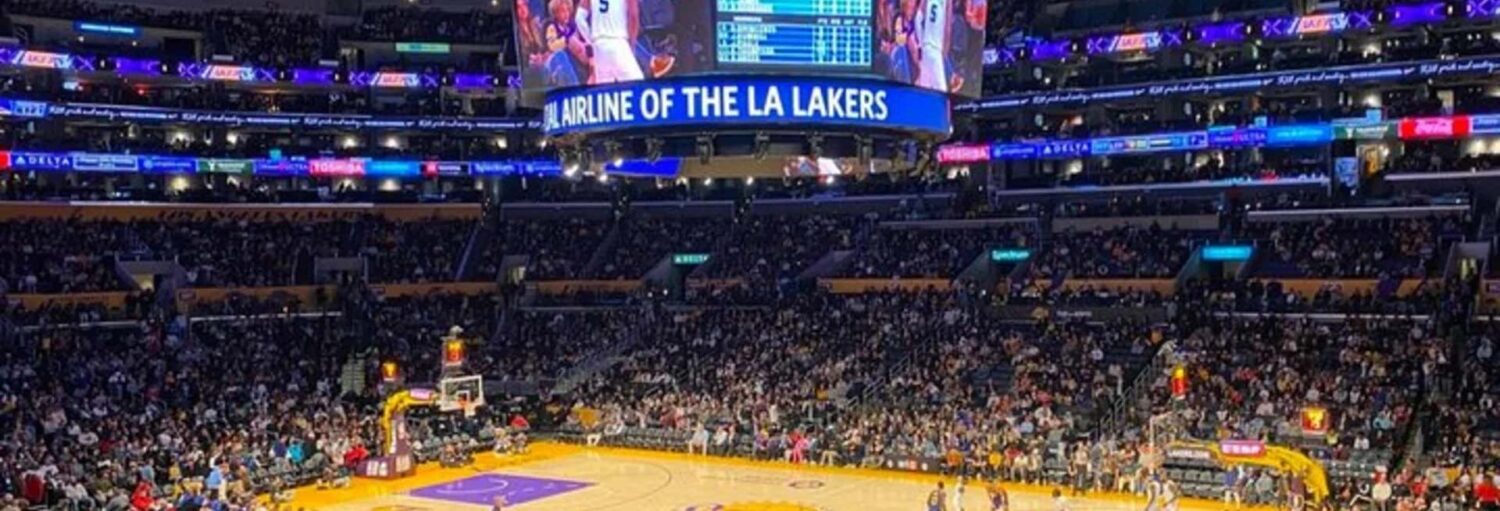 Um jogo de basquete está em andamento em uma grande arena coberta cheia de espectadores, com um placar acima da quadra. A foto é capa do post dos Ingressos da NBA: Los Angeles Lakers.