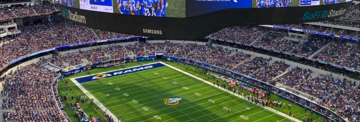 Um estádio de futebol lotado mostra um jogo em andamento no SoFi Stadium com a marca dos Rams visível no campo. A foto é capa do post do Ingresso da NFL Los Angeles Rams ou Chargers.