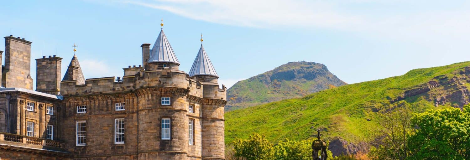 Castelo de pedra com torres cônicas em primeiro plano, encosta verde e céu azul ao fundo. A foto é capa do post do Ingresso do Palácio de Holyrood.