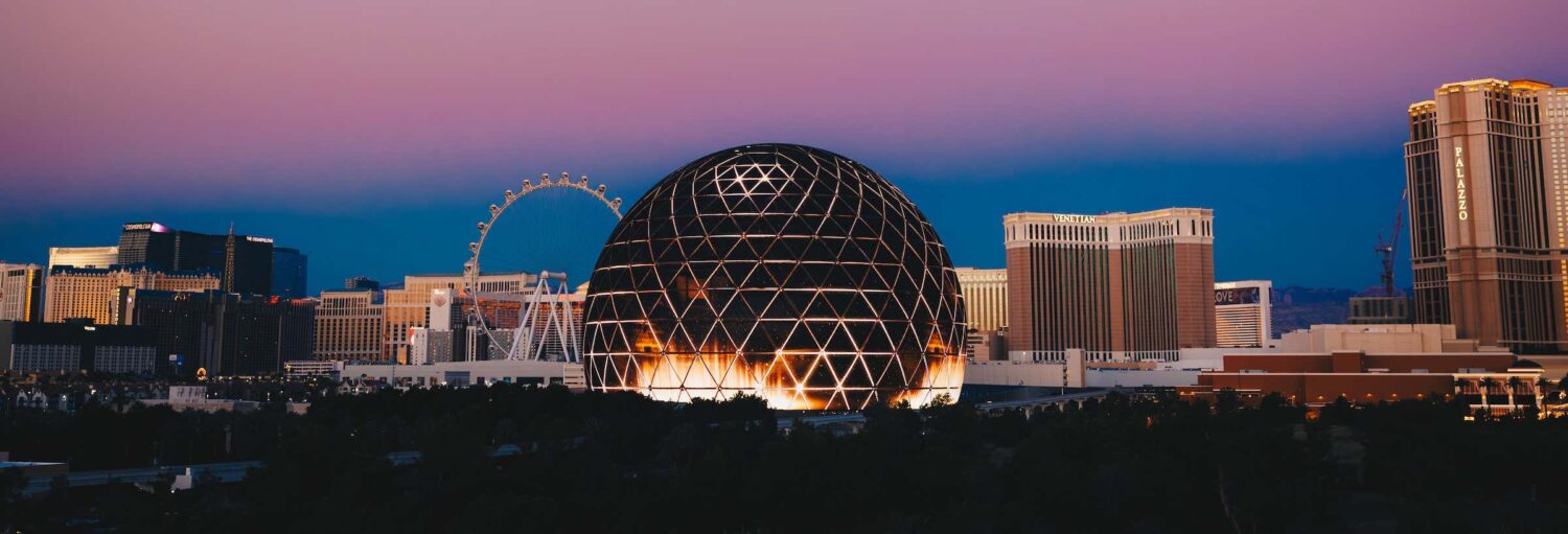 Uma grande estrutura de cúpula iluminada com um padrão geométrico está entre os edifícios ao anoitecer em uma paisagem urbana. Imagem para ilustrar post sobre os ingressos da The Sphere Las Vegas.