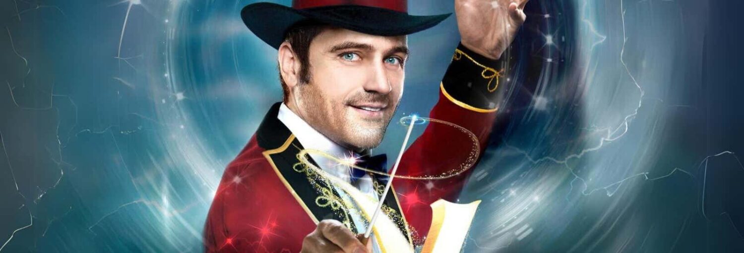 Um homem em um traje vermelho de mestre de circo e cartola preta segura uma batuta e sorri contra um fundo azul e cintilante. Imagem para ilustrar post sobre V - The Ultimate Variety Show.
