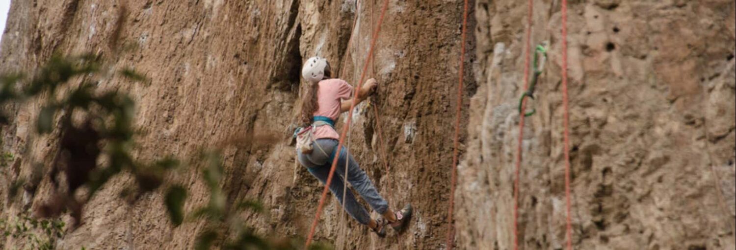 Uma pessoa usando capacete e arnês escala uma rocha íngreme usando cordas.