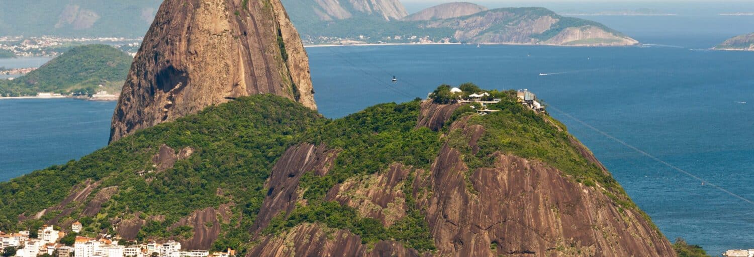 Grandes colinas rochosas com vegetação exuberante à beira-mar, linha de teleférico visível - ideal para a escalada no Pão de Açúcar.
