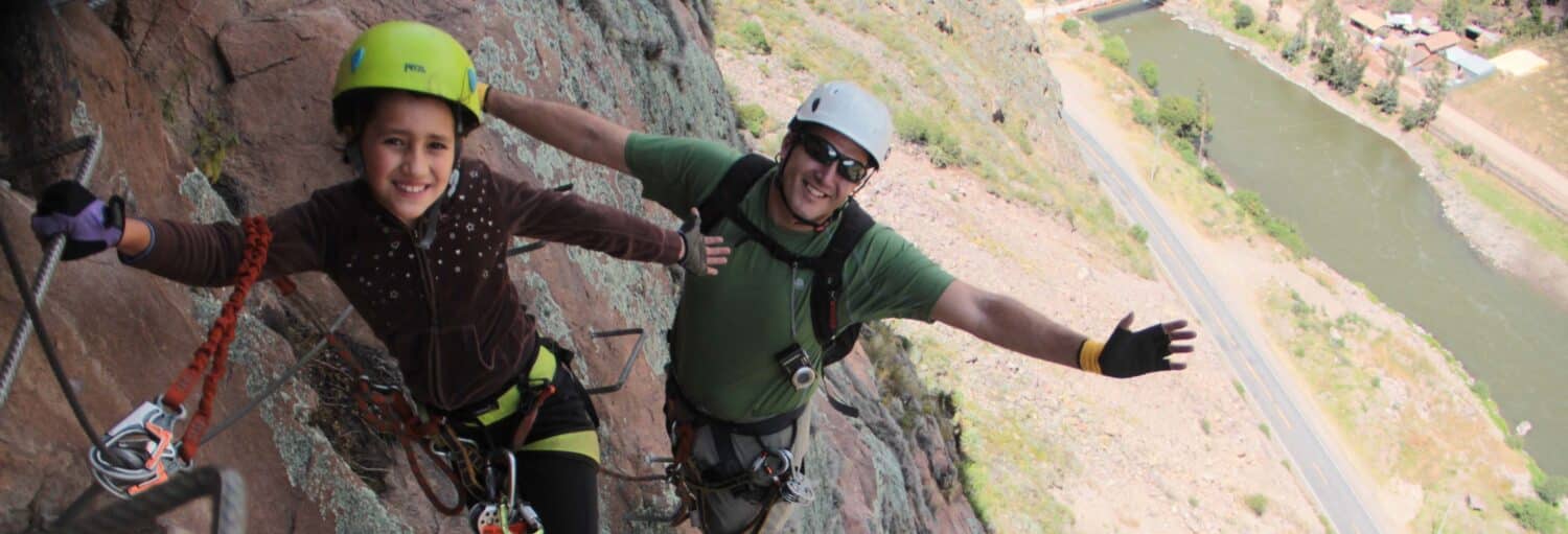 Duas pessoas usando capacetes e arreios escalam uma rocha íngreme, sorrindo e estendendo os braços para fora na Via ferrata do Vale Sagrado dos Incas