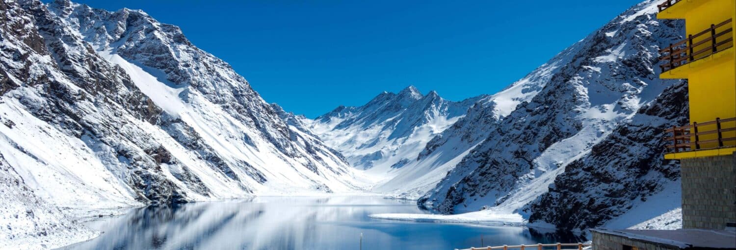 Montanhas cobertas de neve cercam um lago calmo e reflexivo sob um céu azul claro, com um edifício amarelo à direita. A foto é capa do post da Estação de esqui Portillo, Laguna del Inca e Viña San Esteban.