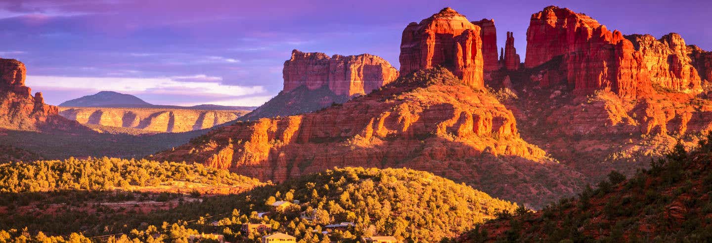 As formações rochosas vermelhas e as mesas são iluminadas pela luz do pôr do sol, com vegetação verde e alguns edifícios em primeiro plano. Imagem para ilustrar post sobre excursão de 3 dias ao deserto do Arizona.