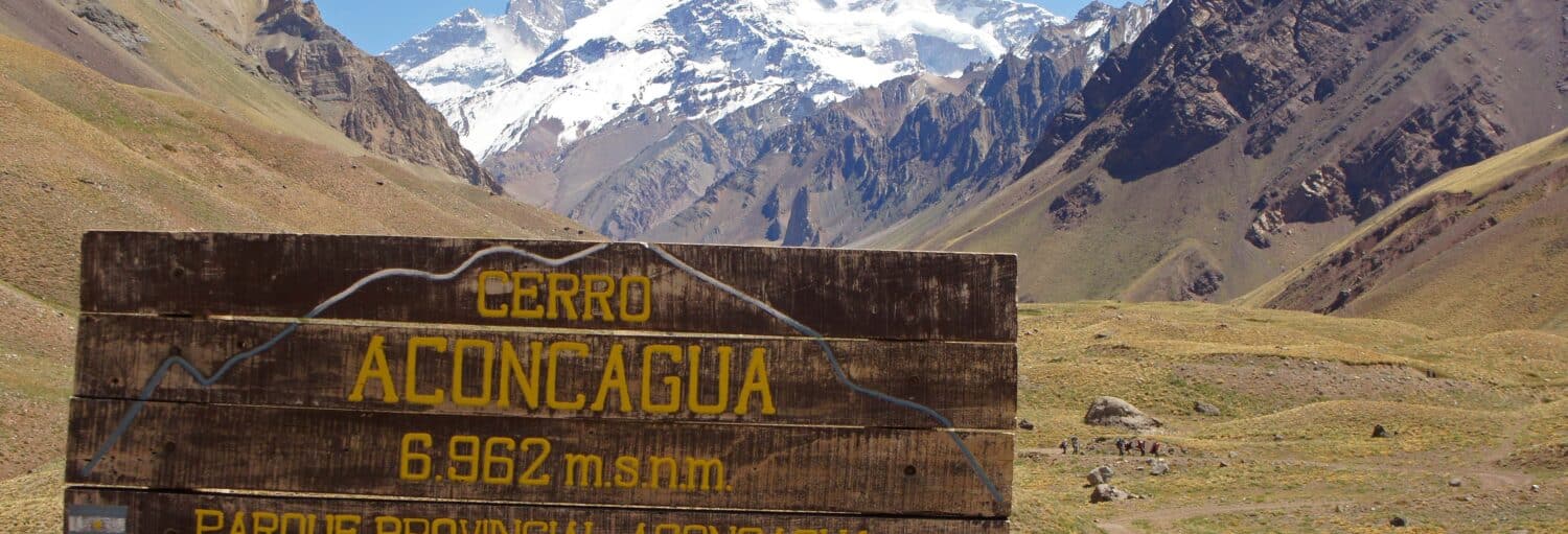 Placa de madeira para o Cerro Aconcágua com montanha coberta de neve e paisagem de vale ao fundo. A foto é capa do post da Excursão ao Parque Aconcagua.