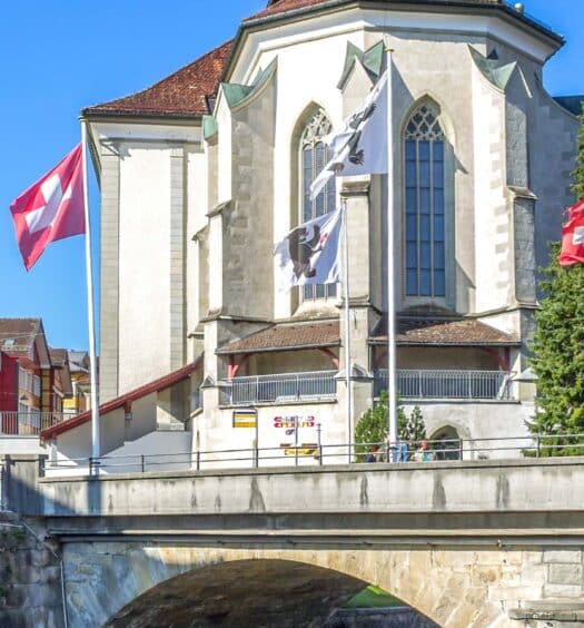 Uma ponte de pedra cruza um rio em frente a uma igreja com bandeiras suíças e edifícios coloridos sob um céu azul claro. Imagem para ilustrar post sobre a excursão a Appenzell e Hoher Kasten.