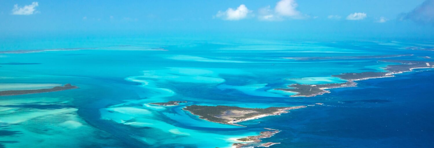 Vista aérea da água azul-turquesa do oceano, ilhas de areia e nuvens espalhadas sob um céu azul brilhante. A imagem ilustra o post sobre Excursão de Miami às Bahamas por conta própria.
