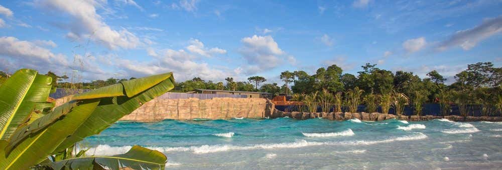 As ondas rolam em uma praia artificial ladeada por palmeiras, com um paredão rochoso e um céu azul ao fundo. - Excursão ao parque aquático Blue Park