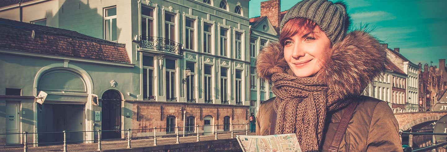 Uma mulher branca, com touca e casaco de pelos sorrindo, posicionada na direita da foto. Atrás dela, há uma rua com prédios de arquitetura medieval - Excursão a Bruges