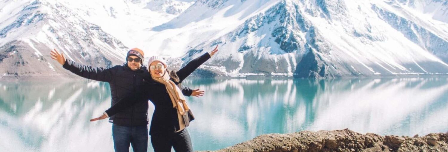 Duas pessoas com roupas de inverno estão de braços dados em um lago, com montanhas cobertas de neve ao fundo. A foto é capa do post da Excursão ao Cajón del Maipo em grupo reduzido.