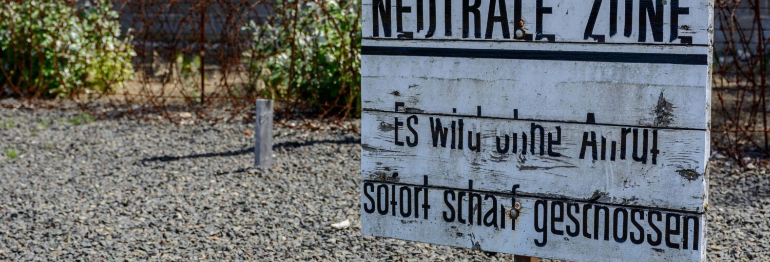 Placa de madeira branca com texto em alemão sobre cascalho, com uma cerca de arame e plantas em um fundo desfocado. Imagem para ilustrar post sobre o campo de concentração de Sachsenhausen.