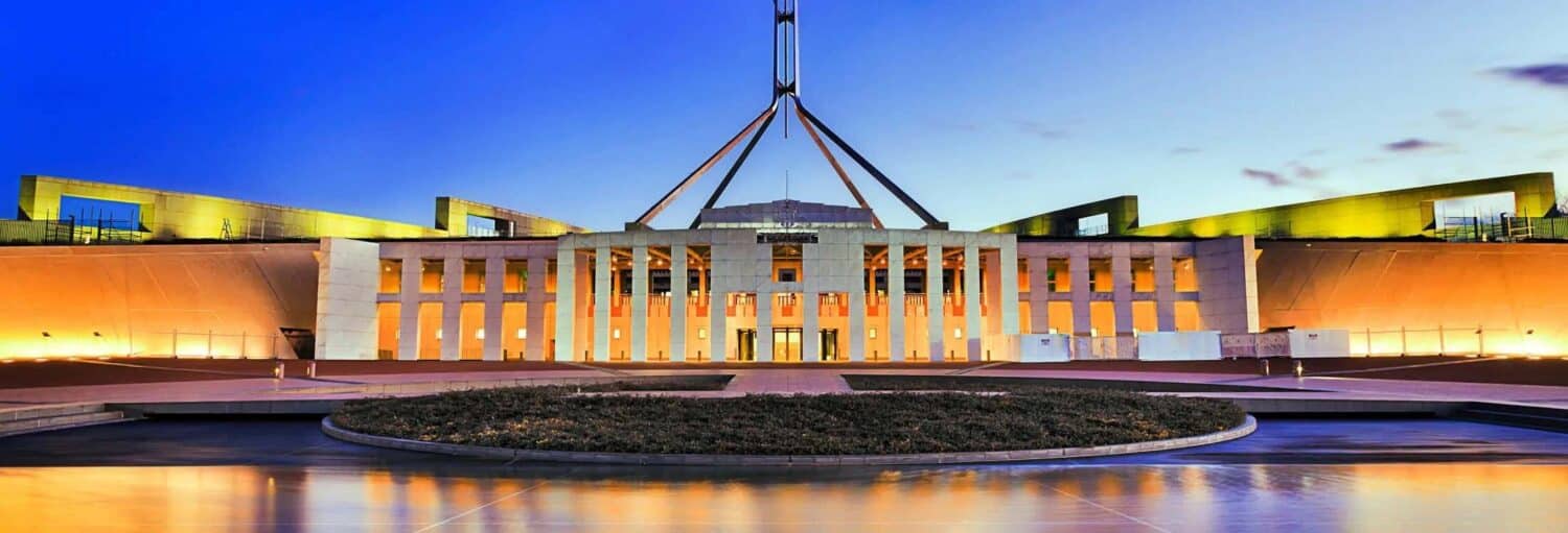 A vista frontal do Parlamento da Austrália em Canberra, iluminado ao anoitecer com um céu azul e amarelo claro. Imagem para ilustrar post sobre a excursão a Camberra.