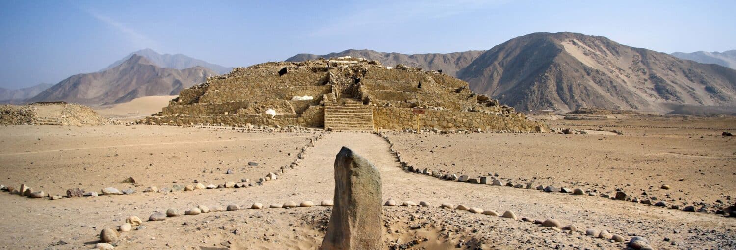 Uma pirâmide de pedra em ruínas em uma paisagem desértica com montanhas ao fundo e um caminho de pedra que leva a ela da Excursão a Caral e Bandurria