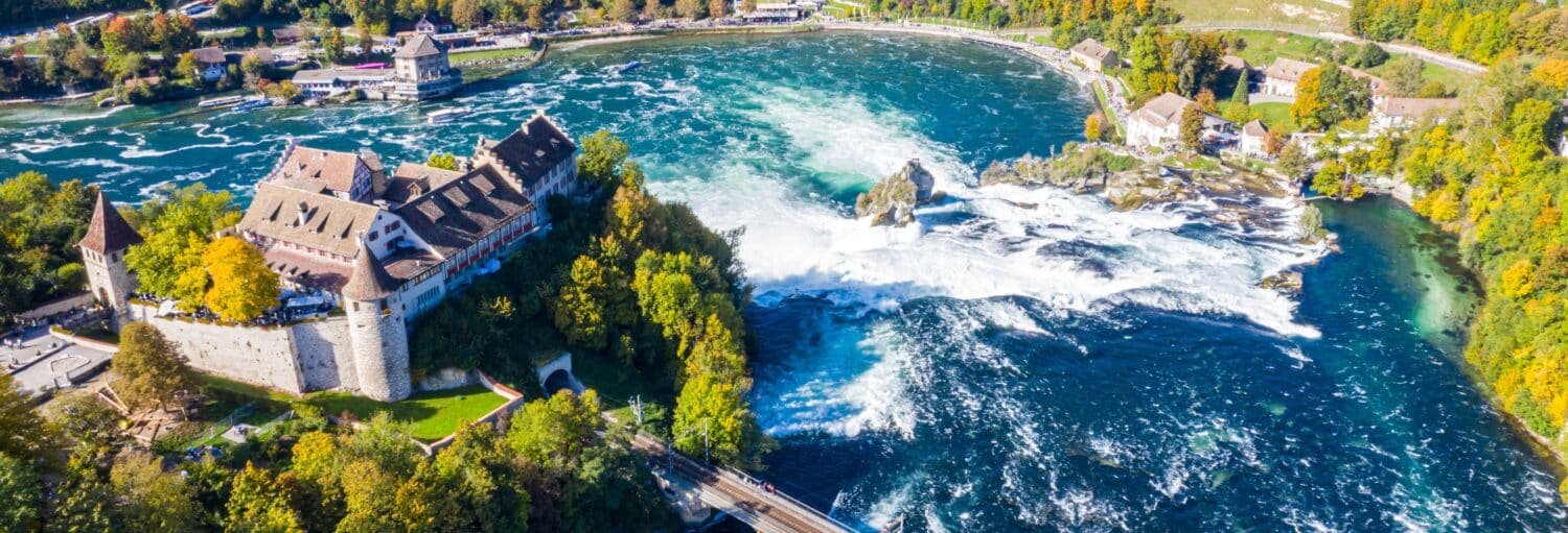 Vista aérea de um grande castelo ao lado de um rio largo com cachoeiras, cercado por árvores e edifícios. Imagem para ilustrar post sobre a excursão às cataratas do rio Reno.