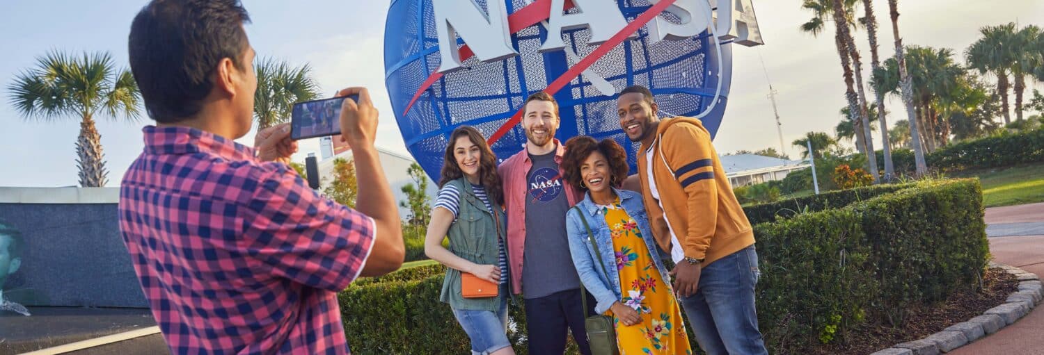 Quatro pessoas posam para uma foto em frente a uma placa da NASA enquanto outra pessoa tira sua foto ao ar livre em um dia ensolarado. A imagem ilustra o post sobre Excursão privada ao Centro Espacial Kennedy.