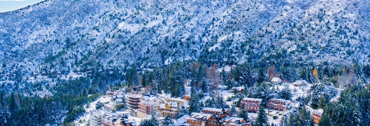 Vilarejo coberto de neve com vários prédios na base de uma encosta de montanha arborizada e coberta de neve na Excursão ao Cerro Catedral