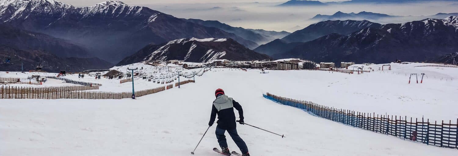 Um esquiador com jaqueta cinza e capacete vermelho desce uma ladeira nevada com montanhas e edifícios ao fundo. A foto é capa do post da Excursão à estação de esqui El Colorado.
