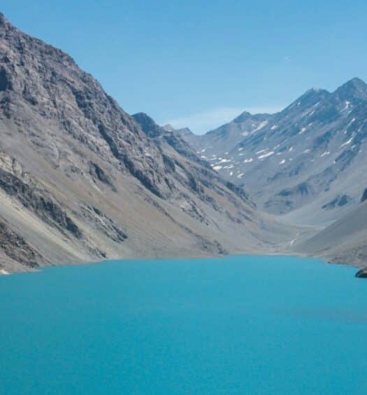 Um lago glacial azul-turquesa é cercado por montanhas íngremes e rochosas sob um céu azul claro. A foto é capa do post da Estação de esqui Portillo e Laguna del Inca.