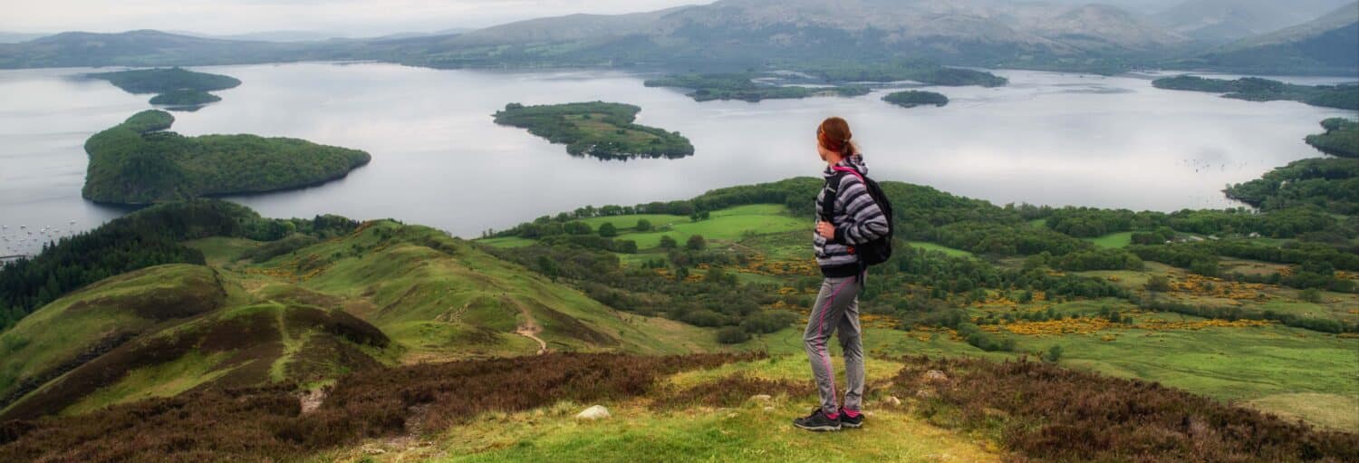 Uma pessoa com uma mochila está em uma colina gramada com vista para um lago com pequenas ilhas e montanhas distantes. A foto é capa do post da Excursão a Glasgow, lagos Lomond e Katrine.