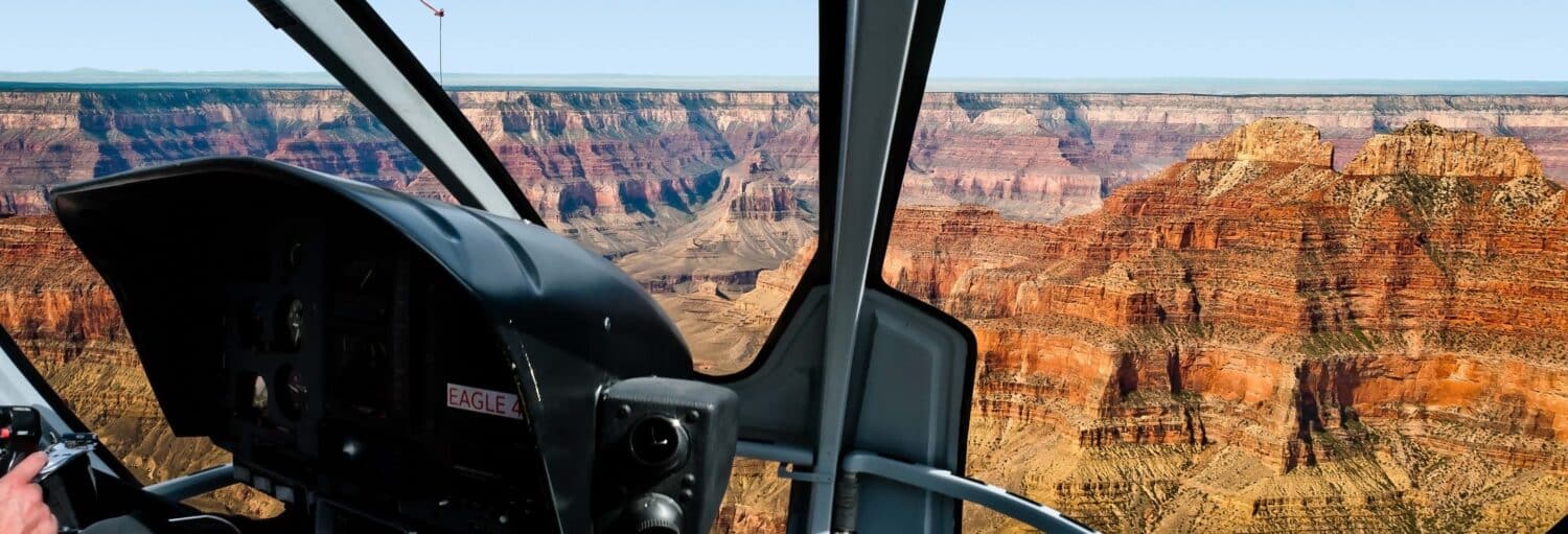Vista do Grand Canyon de dentro da cabine de um helicóptero, com penhascos rochosos e vales profundos visíveis do lado de fora das janelas. Imagem para ilustrar post sobre excursão de helicóptero ao Grand Canyon.