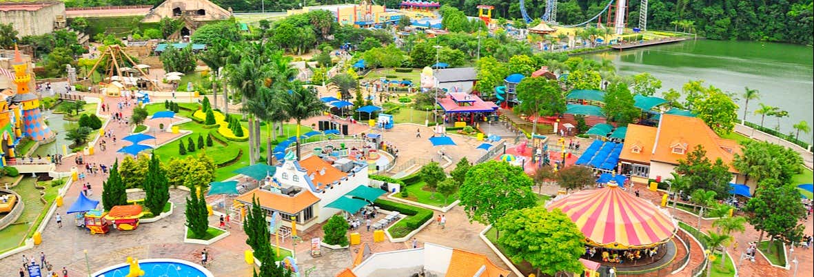 Vista aérea de um parque de diversões com brinquedos, edifícios, árvores verdes, caminhos e um lago ao fundo. A imagem ilustra o post sobre Excursão privada ao Hopi Hari saindo de SP.