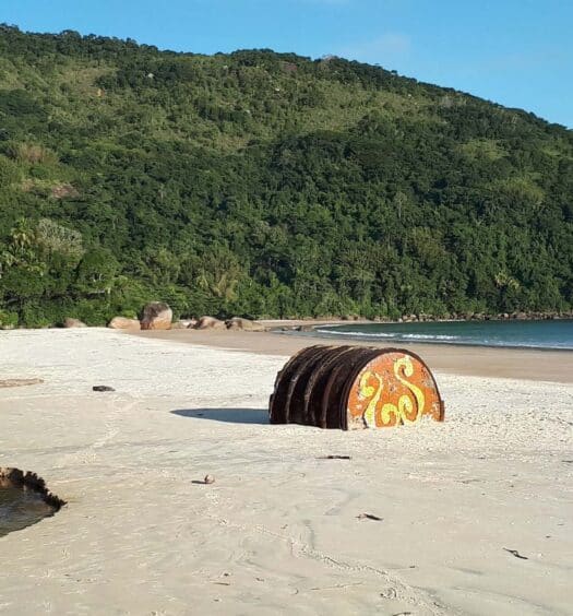 Uma praia de areia em uma trilha pela Ilha Grande no RJ, com colinas verdes e uma escultura de pedra pintada sob um céu claro.