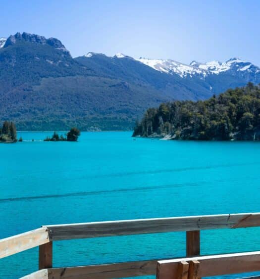 Um lago azul-turquesa claro com ilhas arborizadas e montanhas cobertas de neve ao fundo, visto de uma grade de madeira na Excursão à Ilha Victoria e ao Bosque de Arrayanes