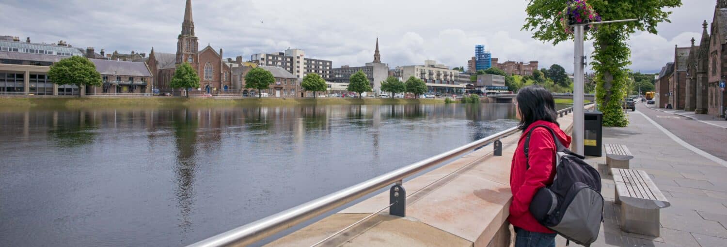 Uma pessoa com uma jaqueta vermelha e uma mochila está à beira de um rio, de frente para prédios e igrejas do outro lado da água. A foto é capa do post da excursão ao Lago Ness, Highlands e Inverness.