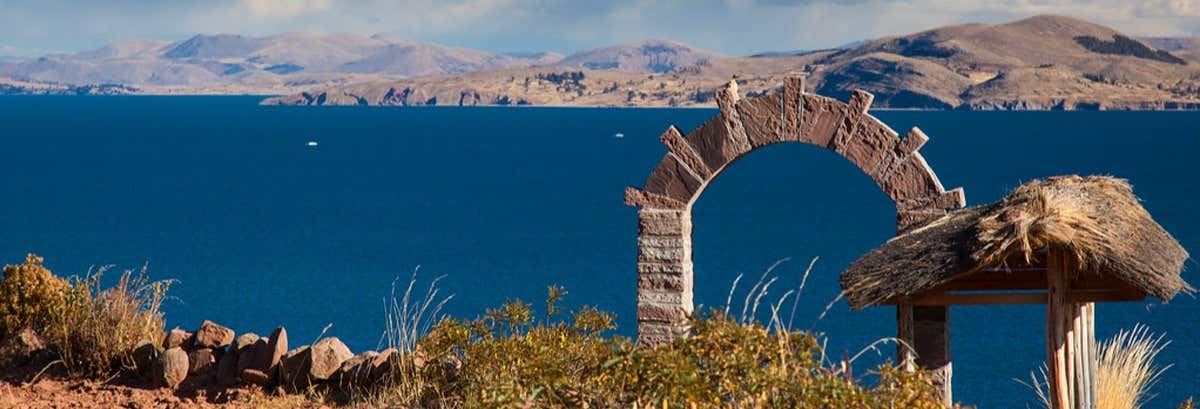 Um arco de pedra e uma estrutura de palha têm vista para um lago azul profundo com montanhas distantes sob um céu parcialmente nublado na Excursão ao lago Titicaca de ônibus noturno