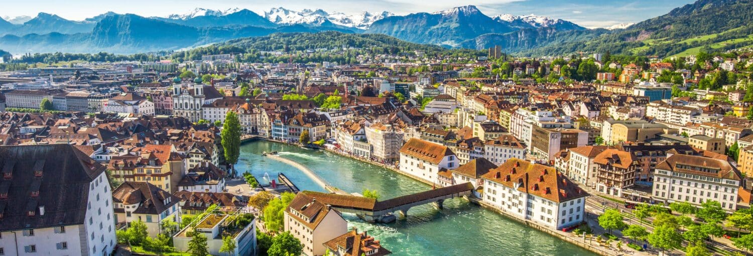 Uma paisagem urbana com um rio, uma ponte histórica de madeira, edifícios com telhados vermelhos e montanhas cobertas de neve ao fundo. Imagem para ilustrar post sobre a excursão a Lucerna.