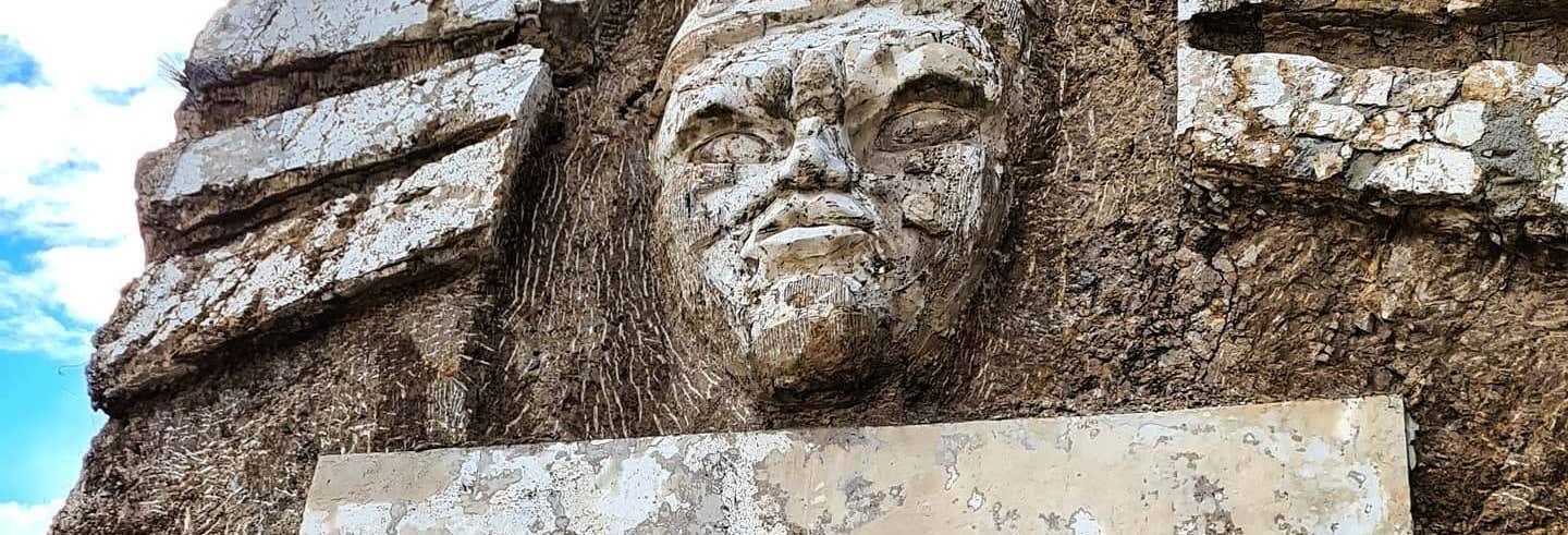 Uma escultura de pedra desgastada de um rosto humano é esculpida em uma superfície de rocha áspera e texturizada ao ar livre na Excursão à Morada dos Deuses