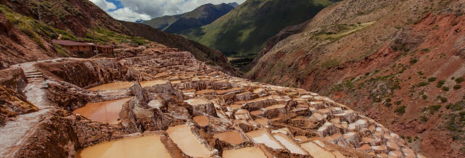 Lagoas de evaporação de sal em terraços na encosta de uma montanha, cercadas por colinas verdes e marrom-avermelhadas sob um céu parcialmente nublado na Excursão a Moray e às salinas de Maras