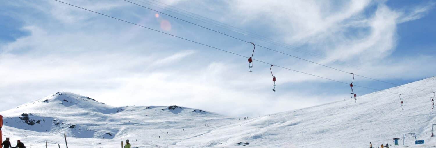 Pista de esqui coberta de neve com um teleférico de superfície e montanhas sob um céu azul com nuvens dispersas. A foto é capa da Excursão ao Parque de Farellones.