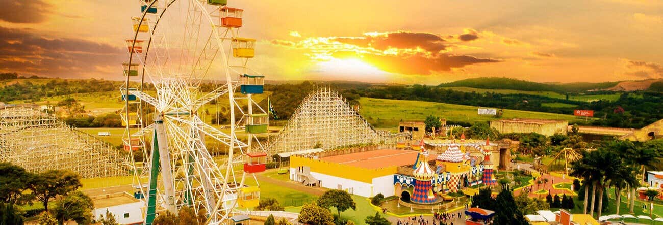 Excursão ao parque Hopi Hari saindo de SP: Roda gigante, montanha-russa e prédios coloridos ao pôr do sol em meio à vegetação.