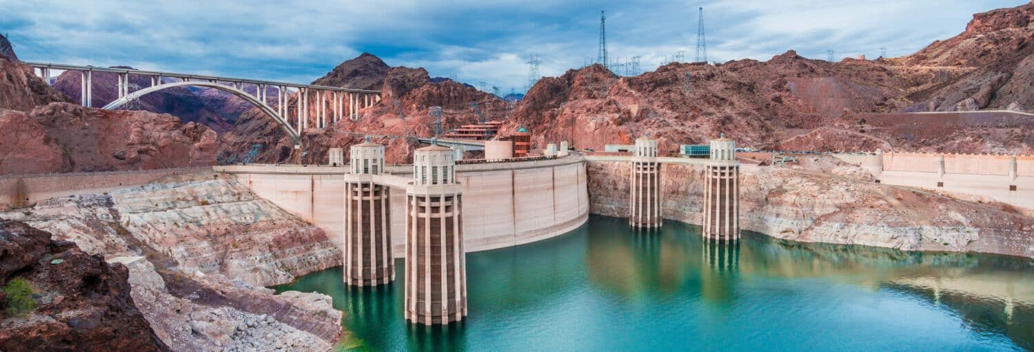 A represa Hoover com torres de entrada e uma ponte, cercada por terreno rochoso e água abaixo da parede da represa. Imagem para ilustrar post sobre excursão à represa Hoover.
