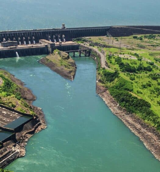 Vista aérea de uma grande represa hidrelétrica com água fluindo entre áreas verdes e infraestrutura próxima.